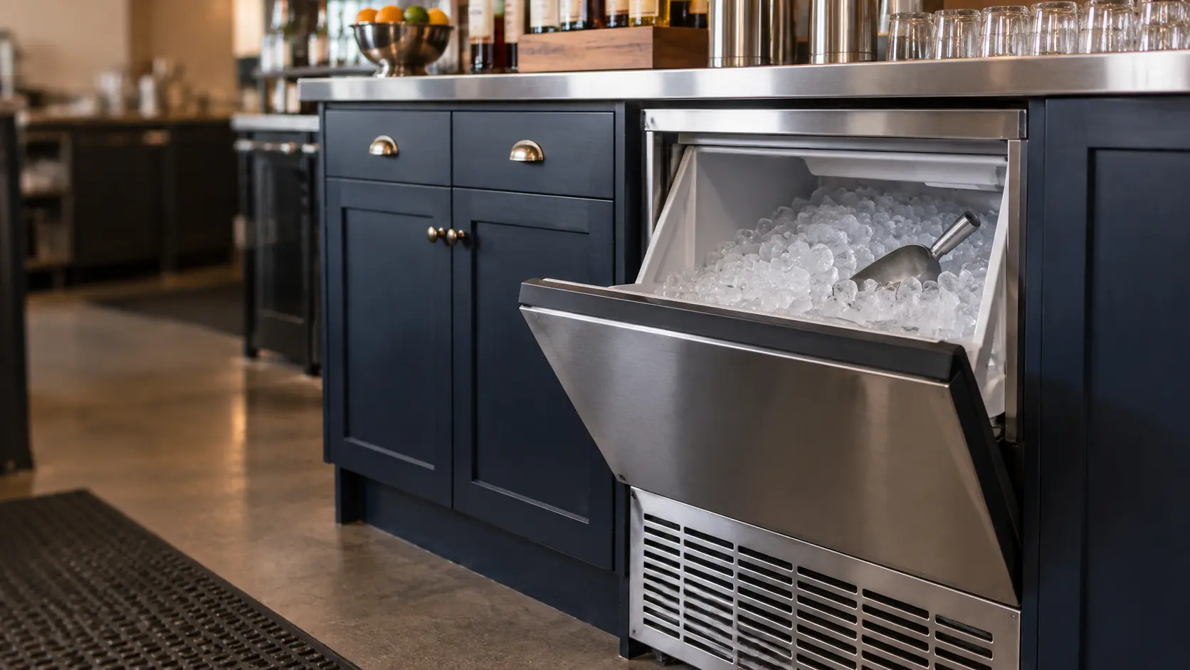 A bar with an undercounter ice machine and navy painted cabinets, seen close at the ice machine's bin door.