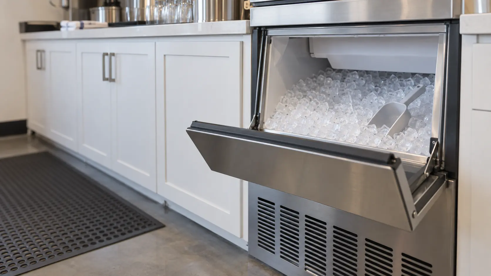 A bar with an undercounter ice machine and white shaker cabinets, seen close at the ice machine's bin door.