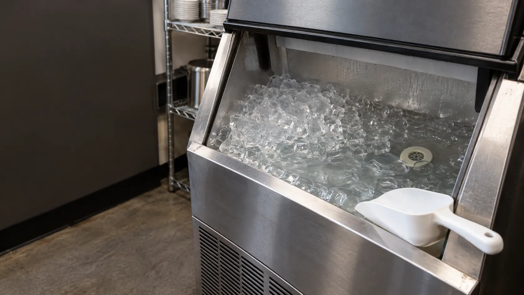 Close view inside the open bin of a stainless ice machine, the pile of clear cubes sitting in a shallow pool of standing water at the bottom of the ice machine's bin, the drain fitting under the water, a white scoop resting on the bin ledge.