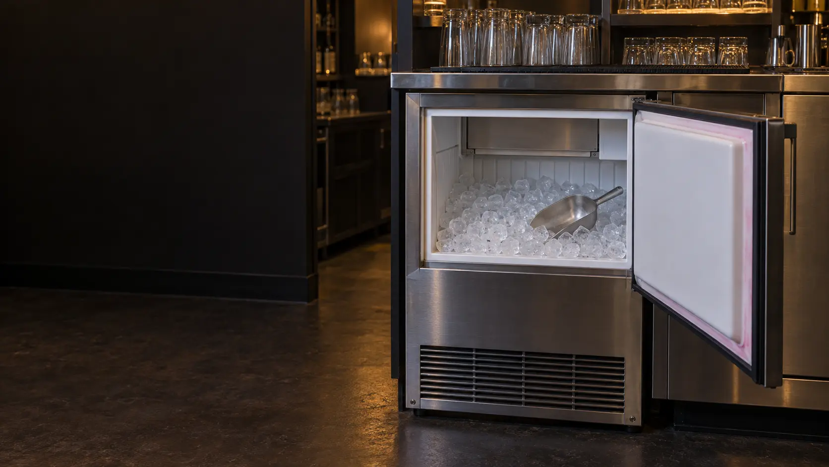 A stainless undercounter ice machine under a dim hotel bar with its insulated door swung open and a scoop of clear cubes resting in the bin, a faint pink film visible along the inside edge of the ice machine door, clean glassware on a shelf above.